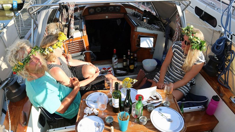 Trio som ska segla jorden runt sitter på midsommaraftonen i sin båt med fat, flaskor och tilltugg, blomsterkransar på huvudet