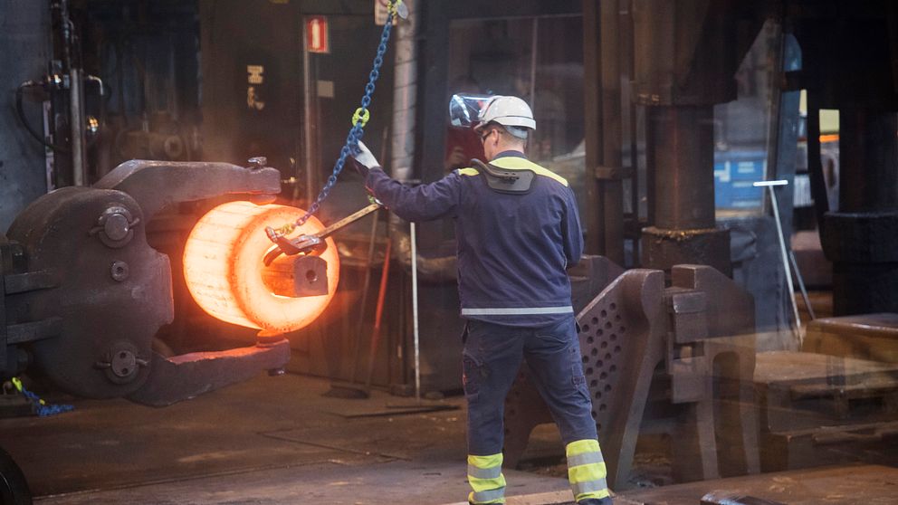 En arbetare i skyddsutrustning står vid en smältugn och hanterar en glödhet metallbit som hänger i en kedja. Den heta metallen avger ett starkt rött sken, och miljön är industriell med fokus på metallbearbetning.