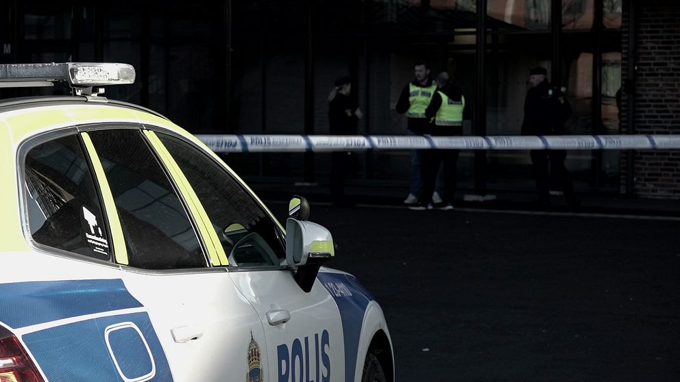 Polisbil parkerad framför avspärrad plats, med poliser i reflexvästar som diskuterar bakom avspärrningstejp.