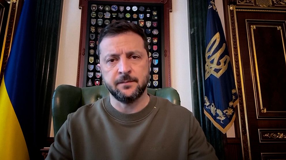 Ukrainas president Volodymyr Zelenskyj sitter vid sitt skrivbord med ukrainska flaggor bakom sig.