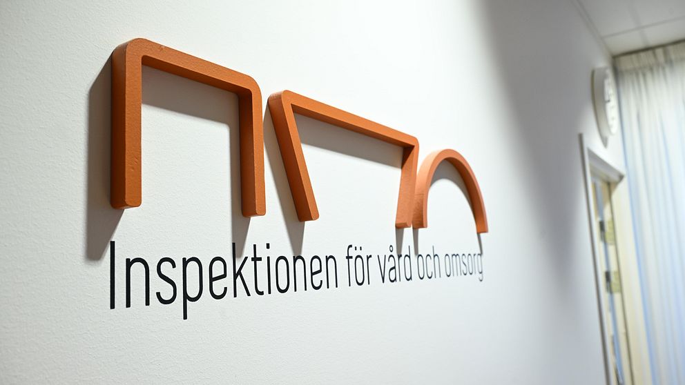 Logotypen för Inspektionen för vård och omsorg, med stiliserade bokstäver i orange och svart, placerad på en vägg i ett kontorsutrymme.