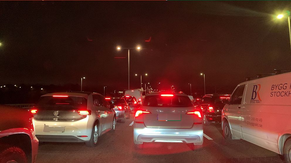 Trafikstockning på en mörk väg, där flera bilar står stilla i kö. Bakljusen lyser rött, och ljus från gatubelysning skapar en dämpad atmosfär. En vit skåpbil med texten ”BYGG STOCKHOLM” syns till höger.