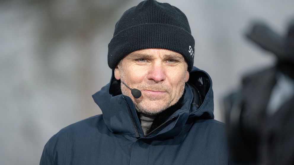 Person i mörk vinterjacka och mössa utomhus, träd syns suddigt i bakgrunden.