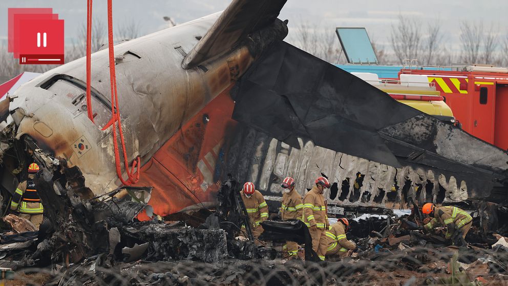 Brandmän inspekterar vraket av ett kraschade flygplan som har tagit skador av brand, med omsorgsfullt arbete i närheten av det förstörda fordonet. En rad räddningstjänstfordon syns i bakgrunden.