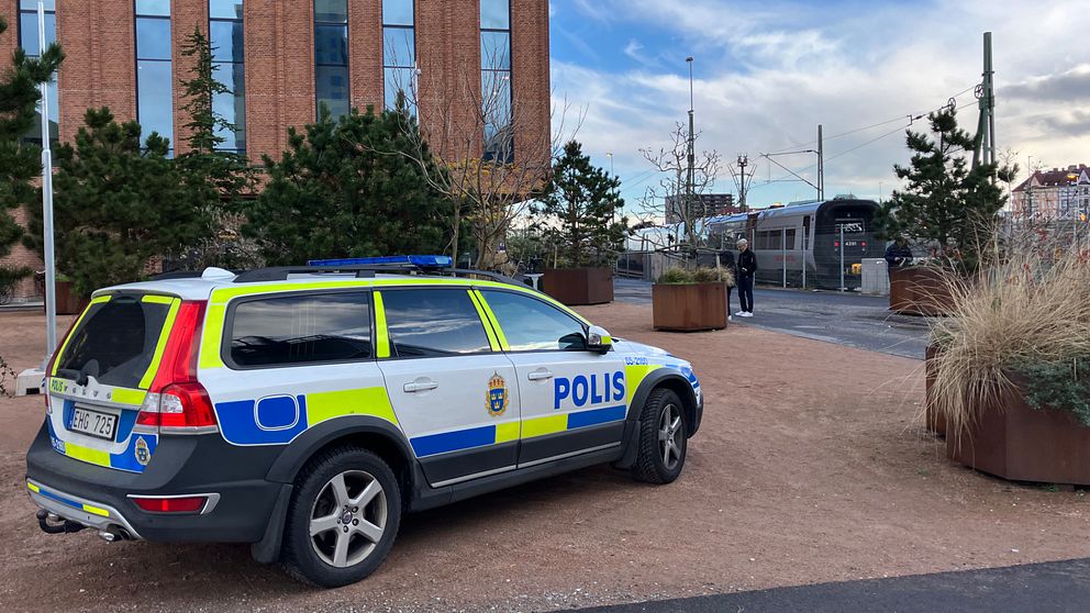 En polisbil parkerad i en urban miljö med ett modernt byggande i bakgrunden. Området är prytt med träd och växtlighet, och en person syns längre bort i bilden, vid en väg eller spår.
