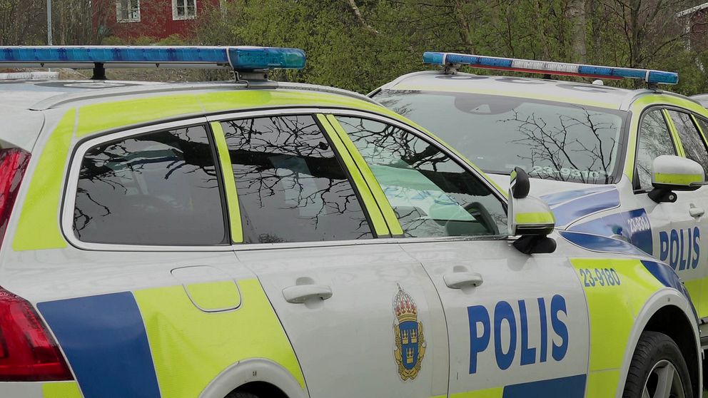 Två polisfordon står parkerade med tydlig synlighet av bilarna och deras blåljus, omgivna av träd i bakgrunden.