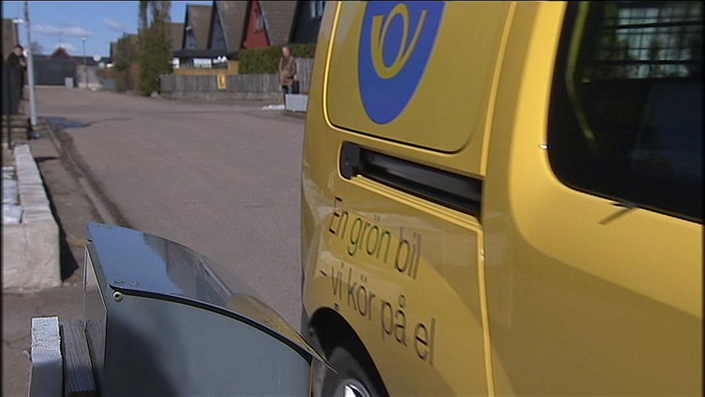 En elbil i gult står parkerad vid en väg. I bakgrunden syns en asfaltgata med hus längs sidan. På bilen finns texten ”En grön bil – vi kör på el”. En grå postlåda syns i förgrunden.