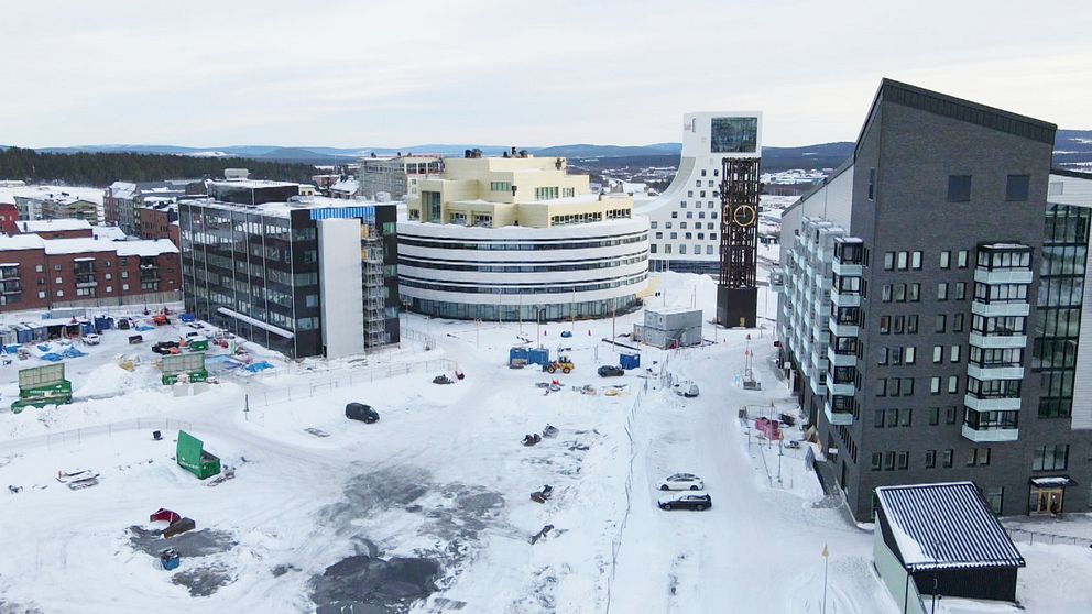 Vy över en snöklädd byggarbetsplats med flera moderna byggnader i en stadsmiljö. Det är delar av Kirunas nya centrum. I mitten syns en stor, rundad byggnad med en ljus färg, medan omgivande byggnader har en mer traditionell arkitektur. Snön täcker marken och det finns byggmaskiner samt annat arbetsmaterial synligt. I bakgrunden kan man se fler stadsbyggnader och en vidsträckt himmel.