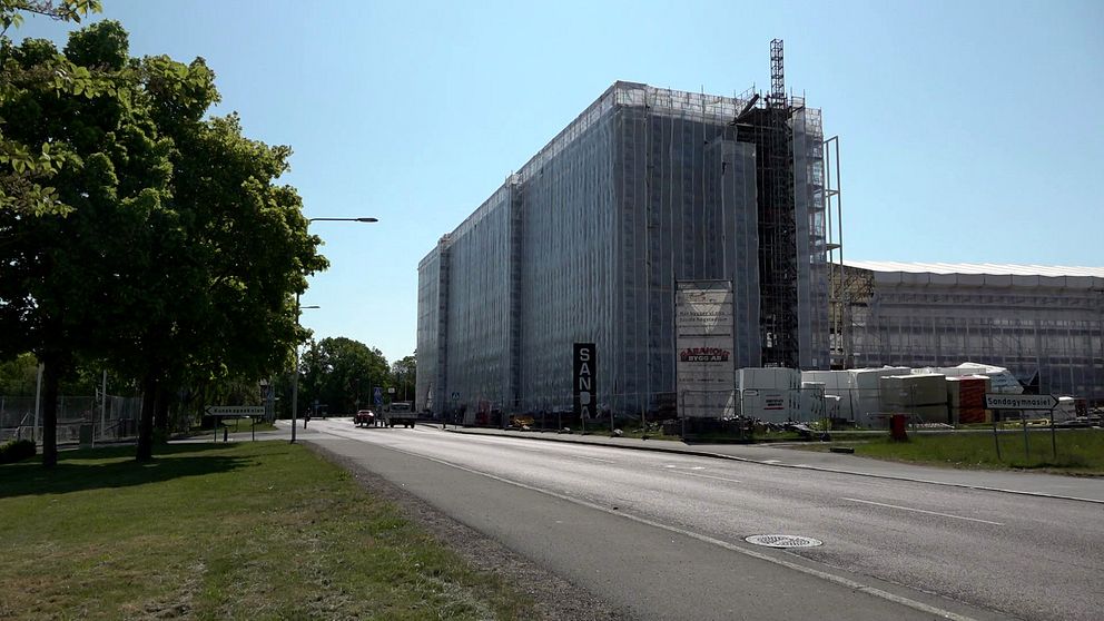 Byggarbetsplats med en hög byggnad som är täckt av skyddande plast och stillastående byggnadsställningar, fotograferad från en väg i dagsljus. Träd syns till vänster om bilden, och det finns byggmaterial och en skylt i närheten.