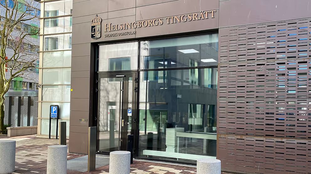 ALT-text: Ingången till Helsingborgs tingsrätt med en modern glasfasad och synlig skylt. Området kring entrén har stensatta gångar och parkeringsplatser. Träd och moderna byggnader syns i bakgrunden.