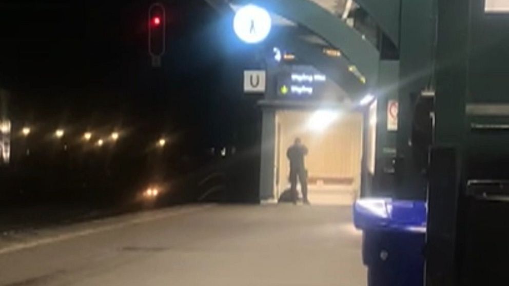 En nattbild av en polis som står på en upplyst tågplattform, med ett tåg som närmar sig i fjärran.