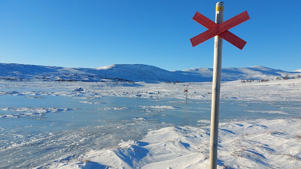 Ett snötäckt och isigt landskap under en klarblå himmel, med ett rött kryssmärkt ledkryss i förgrunden. Fjäll syns i bakgrunden.