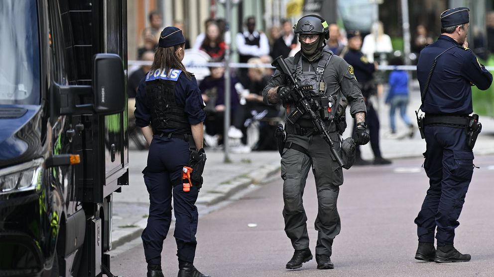 Poliser står på en gata där en incident har inträffat. En beväpnad polis i taktisk utrustning går framåt, medan en annan polis övervakar situationen. I bakgrunden syns en folkmassa och ett polisfordon. Området är avspärrat med avspärrningsband.