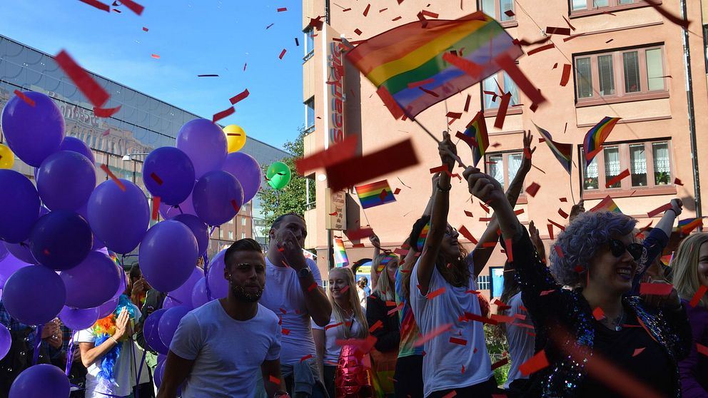 Personer firar Pride med regnbågsflaggor och lila ballonger. Confetti i luften.