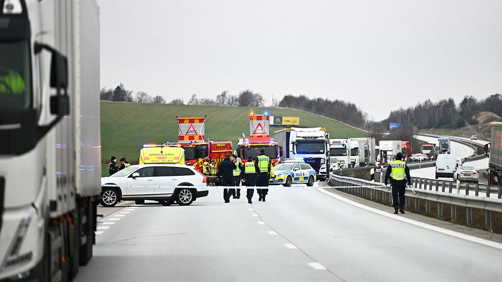 En motorväg som blockerats av räddningstjänst och polis efter endöd man påträffats i höjd med Skrea. Flera fordon, inklusive lastbilar och ambulans, syns i bakgrunden. Räddningsteam arbetar på platsen, medan poliser dirigerar trafiken. Målet med insatsen tycks vara att hantera olyckan och säkerställa trafiksäkerheten.