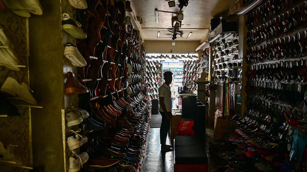 En man står inne i en mörkt belyst skobutik med hundratals skor prydligt uppställda på hyllor längs väggarna. Han är belyst bakifrån av ljuset från den öppna dörren i butikens bakre del.