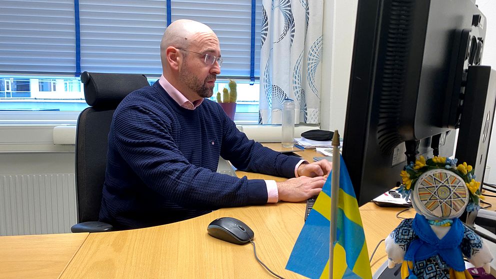 Timur Uman, professor i företagsekonomi vid högskolan i Jönköping, sitter vid ett skrivbord i ett kontorslandskap, klädd i en blå tröja. Han använder en dator med en stor skärm. På skrivbordet finns en mus och ett par små svenska flaggor. I förgrunden syns även en dekoration med traditionell svensk klädsel. Rummet är upplyst av naturligt ljus som kommer från fönstren bakom honom.