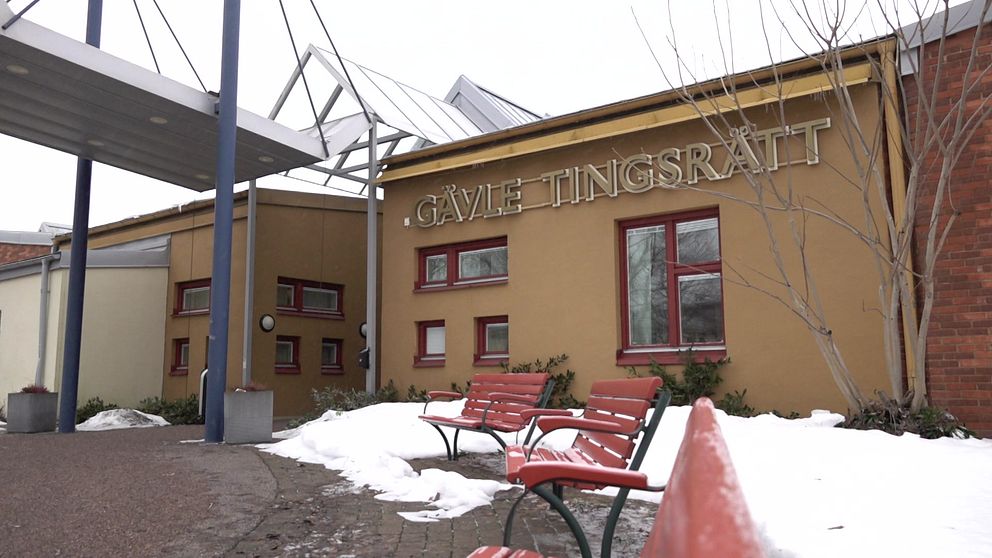 Utomhusbild av Gävle Tingsrätts byggnad på vintern med snö på marken och röda parkbänkar i förgrunden.
