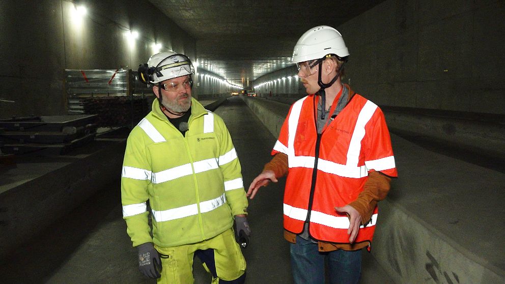 Jimy Andersson, byggledare för Västlänken vid centralstationen i Göteborg, till vänster i en grön reflexjacka. Carl Malmborg, reporter för SVT Nyheter Väst, till höger i orange reflexväst. De båda går och pratar i en lång tunnel som är en del av Västlänken.
