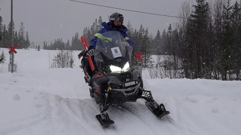 Person kör snöskoter i vinterlandskap med snöklädda träd i bakgrunden.