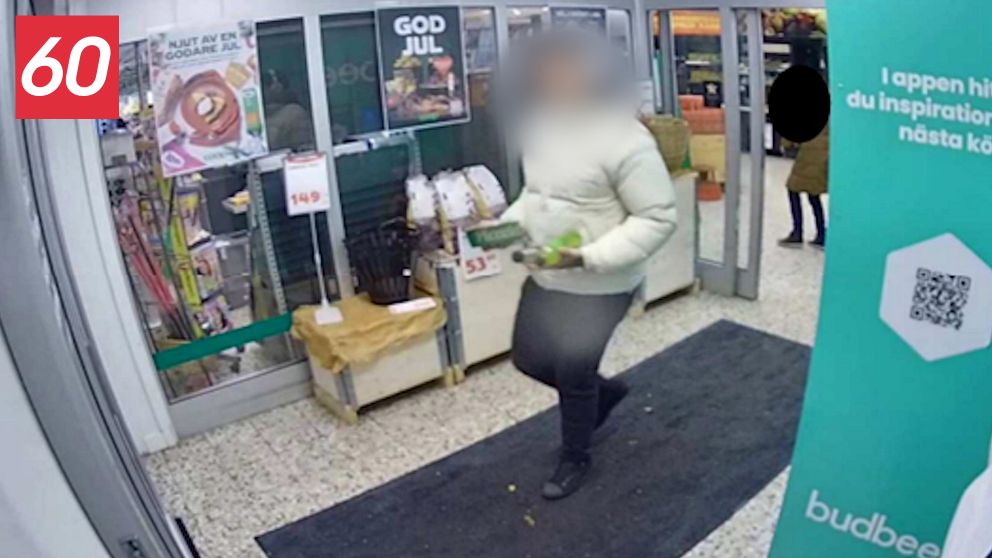 En person i vit tröja och mörka byxor som bär på en påse med grönsaker, går ut genom entrén till en butik. I bakgrunden syns affischer med julhälsningar och en skylt som annonserar olika produkter. Det finns även en grön väggreklam för en applikation till höger om bilden.