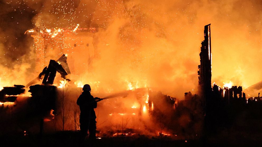 En brandman sprutande vatten på en intensiv brand som har gripit tag i en byggnad. Flammor och rök fyller luften, och omgivningen är upplyst av den starka elden. I bakgrunden syns förvridna och brinnande trästrukturer.