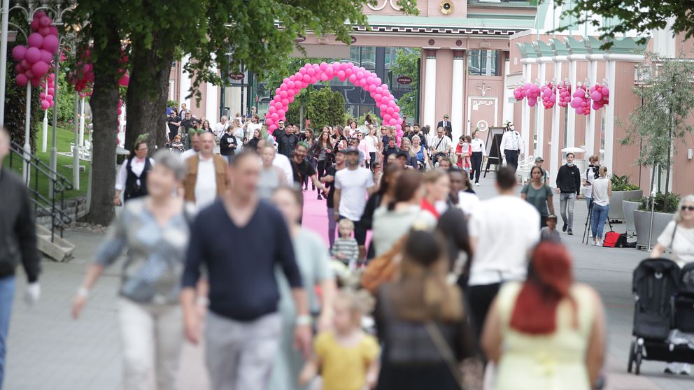 Folkmassa passerar under rosa båge i stadsmiljö med grönska och blomdekorationer.