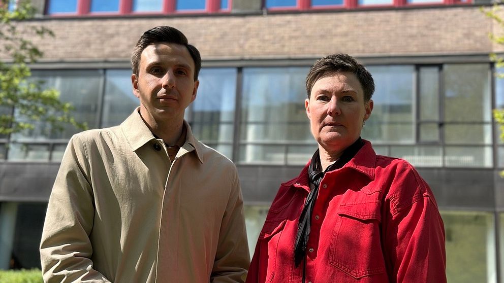 Två personer står framför en byggnad med stora fönster och rödbruna fönsterkarmar.