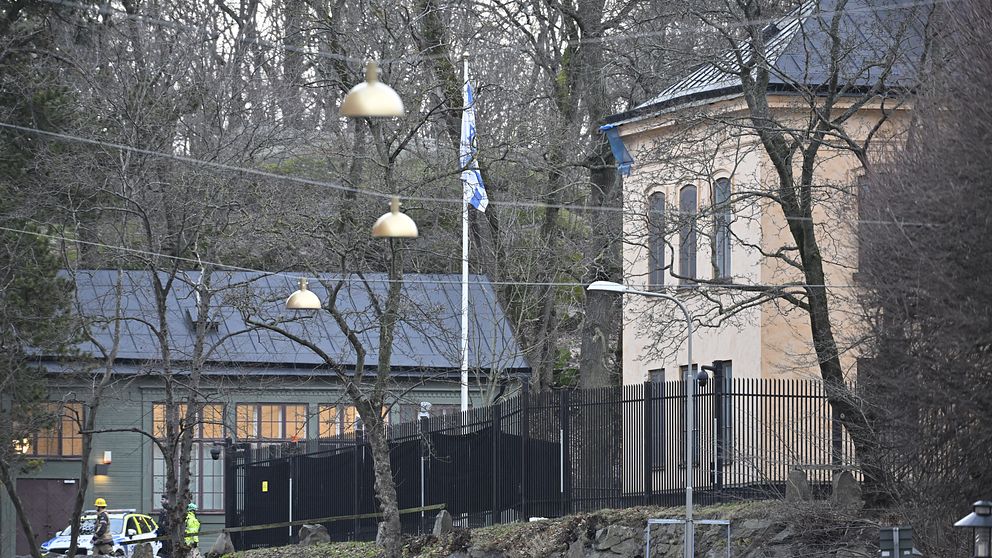 Byggnad med flaggstång omgiven av träd, en person i reflexkläder syns i förgrunden.