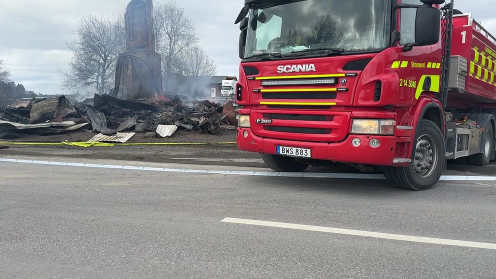 Brandbil framför ruiner efter en brand, rök syns från resterna i bakgrunden.