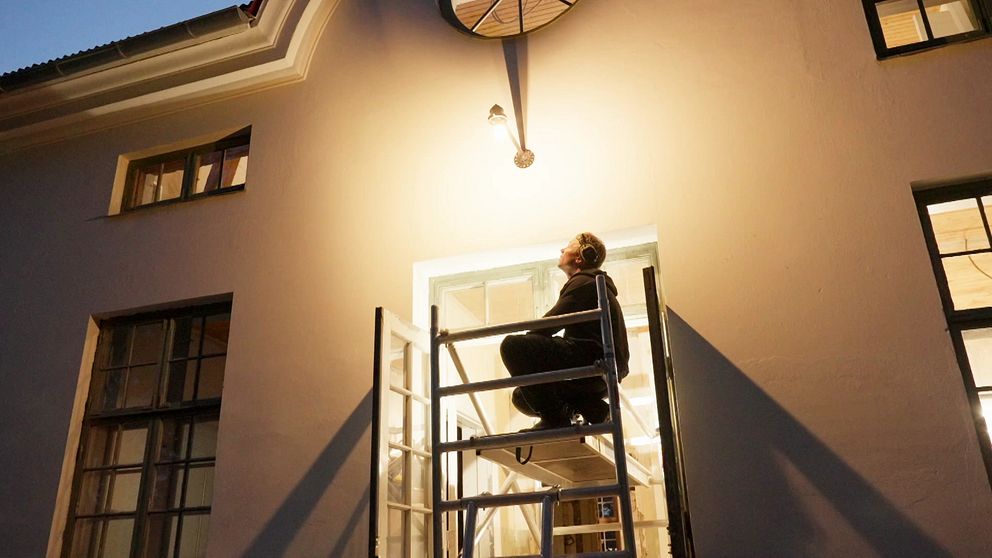 Person på byggställning monterar lampa över entré i skymning.