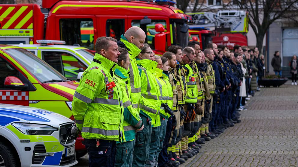 Räddningspersonal och poliser står på rad framför en rad utryckningsfordon, inklusive ambulanser och brandbilar, under en utomhusceremoni eller samling.