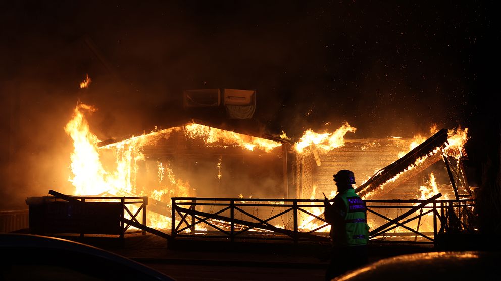 En byggnad står i lågor på natten, en brandman syns i förgrunden.