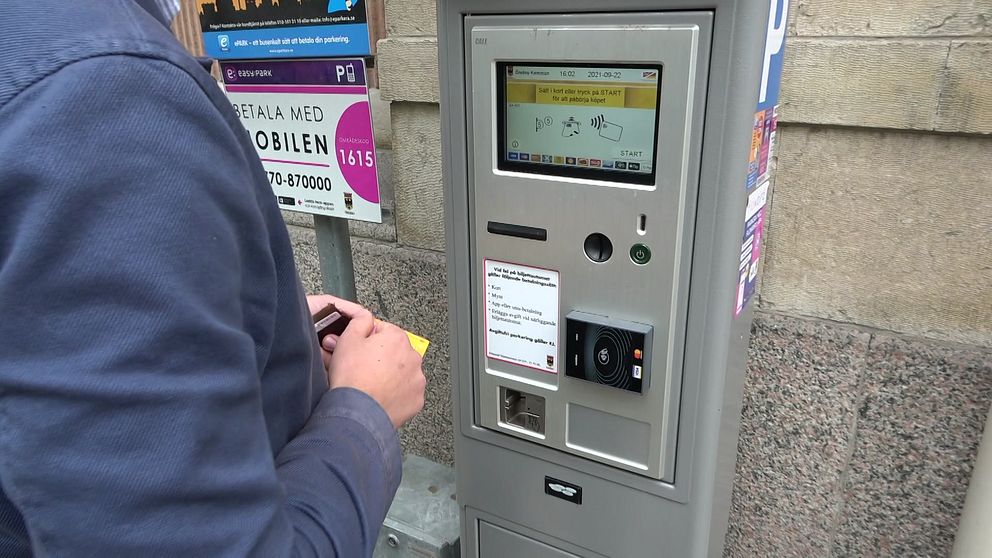 En person står vid en automat för betalning av parkering, där de håller en biljett eller kort i handen. Automaten har en digital skärm som visar olika options för betalning och instruktioner. I bakgrunden syns en informationsskylt om parkering.