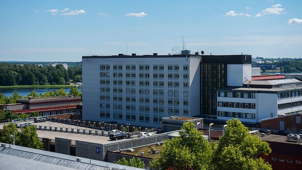 Sjukhusbyggnad omgiven av träd och en sjö i bakgrunden under en klarblå himmel.