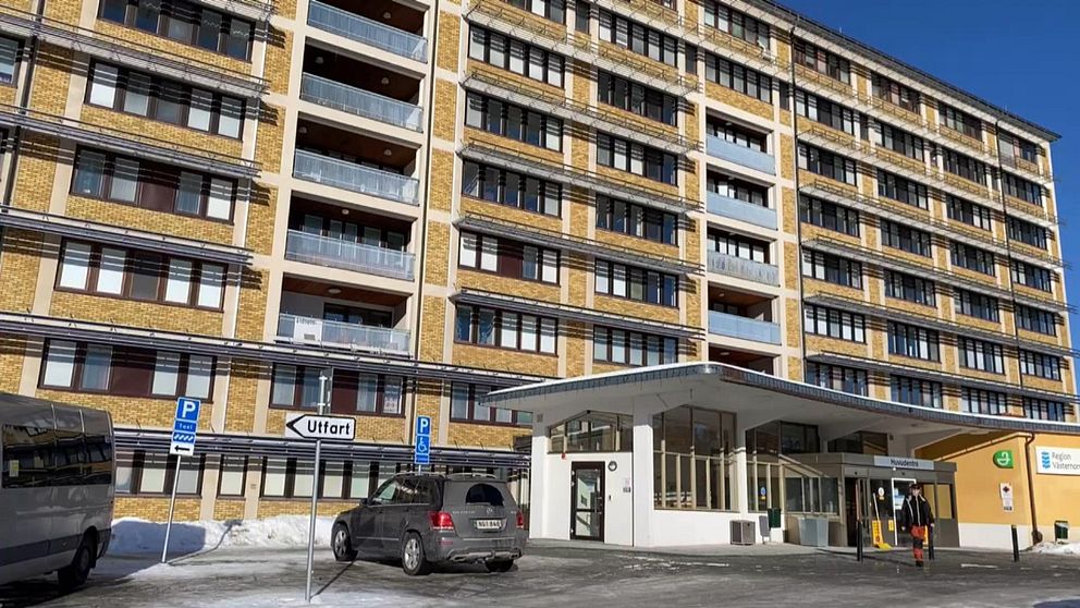 Entrén till ett sjukhus med parkerade bilar utanför och ett flervåningshus i bakgrunden.