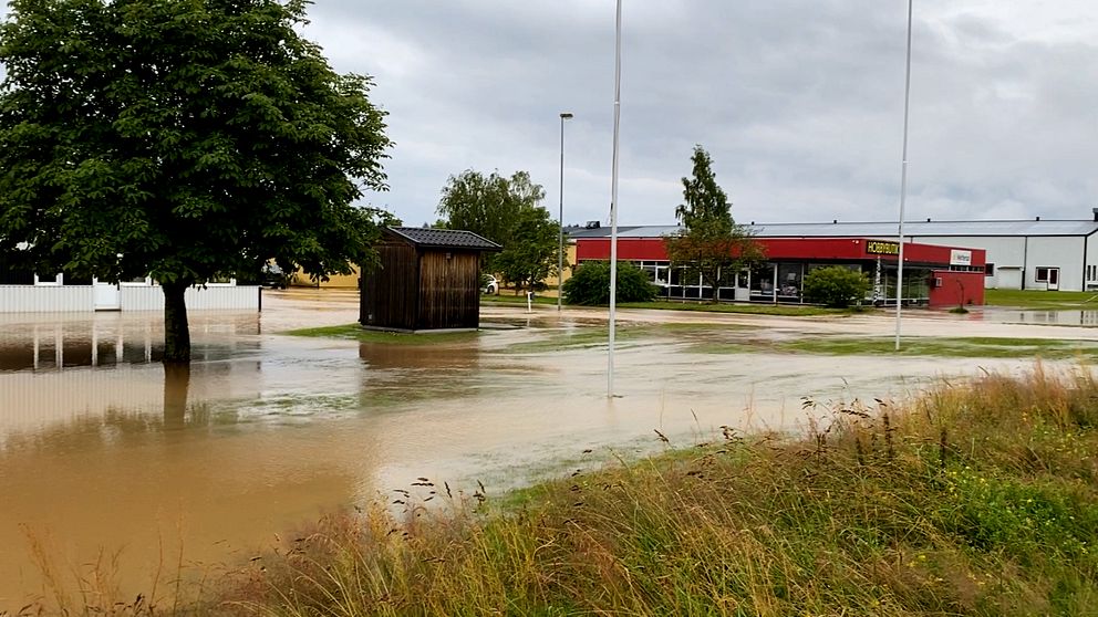 Översvämmad gata vid industribyggnad, gräsfält och träd under molnig himmel.