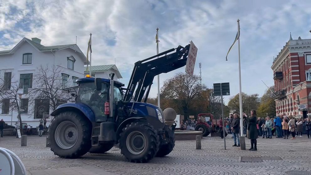 En blå traktor med en lyftanordning framför en folkmassa som samlats på en torg. Bakgrunden visar både moderna och äldre byggnader samt flera flaggstänger med flaggor. Traktorn bär ett skylt som inte är tydligt synlig, och det är en klar himmel med några moln.