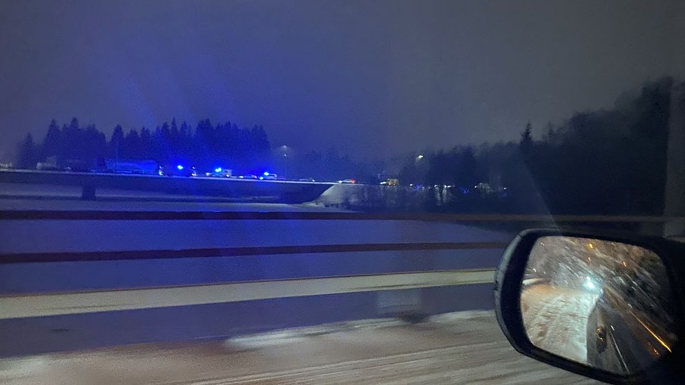 Nattbild tagen från en bil som visar en bro med blåa blinkande ljus från ett räddningsfordon på sidan av vägen, omgiven av snöiga omständigheter och mörka träd i bakgrunden.