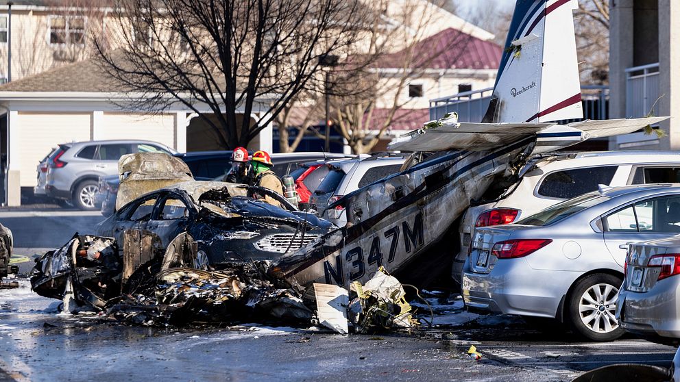 Litet flygplan kraschat på parkering, omgiven av brandskadade bilar. Räddningspersonal inspekterar.