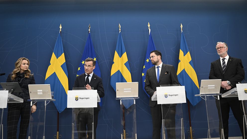 En kvinna och tre män i kostymer håller en pressträff framför svenska flaggor och blå bakgrund.