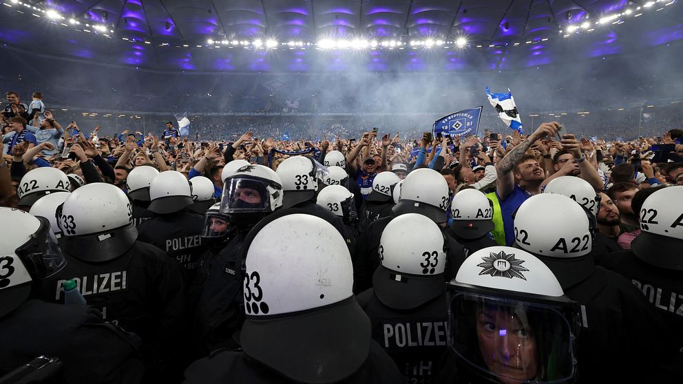 Poliser i hjälmar bildar kedja framför stor fotbollspublik på en upplyst arena.