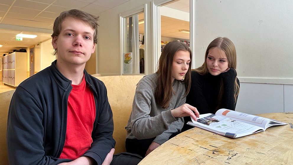 Tre personer sitter runt ett bord och tittar i en skolbok. En ljus korridor sträcker sig i bakgrunden.