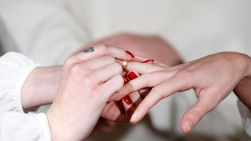 Två händer är synliga där en person sätter en ring på den andra personens finger. Den som sätter på ringen har naglar målade i röd färg, medan den andra handen har en ljusare nyans och är prydd med en enkel ring. Bakgrunden är suddig och antyder en ceremoniell eller romantisk atmosfär.