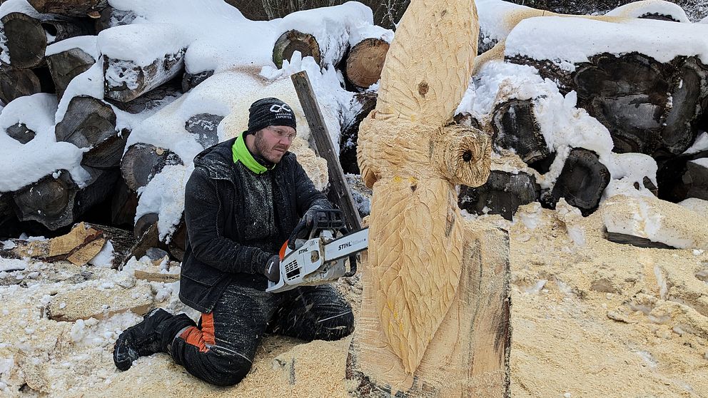 Motorsågsskulptören Johan Karlsson sitter på knä med en motorsåg i händerna och formar en stor trästock till en uggla