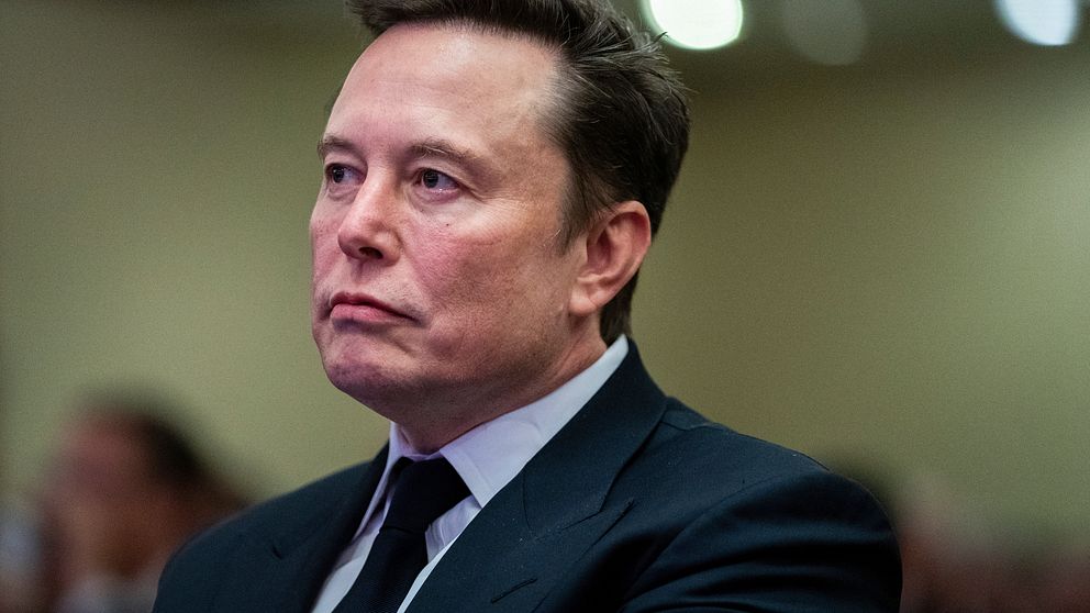 Elon Musk i en mörk kostym sitter i ett möte eller en offentlig sammanhang. Bakgrunden har suddiga detaljer som tyder på en inomhusmiljö med mjukt ljus och oskarpa personer i närheten.