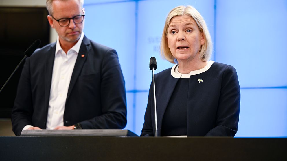 Två personer står vid ett podium under en presskonferens med en digital skärm i bakgrunden.