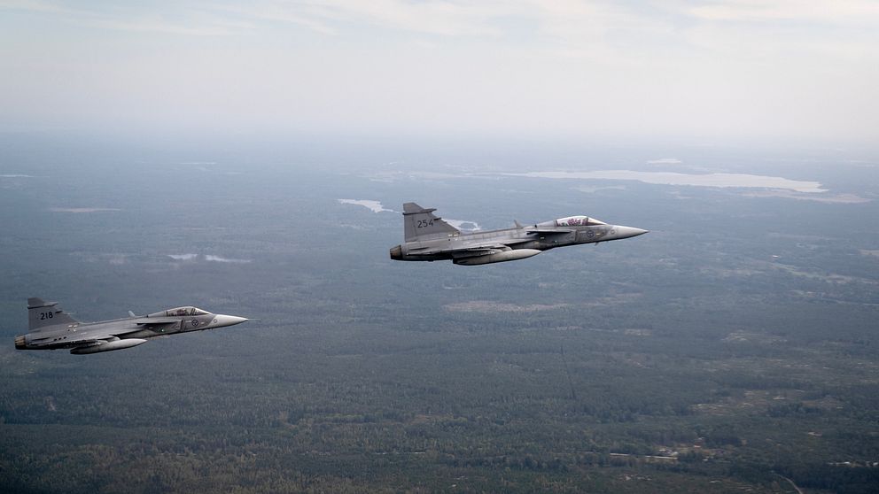 Två stridsflygplan i formation flyger över ett landskap med skog och sjöar.