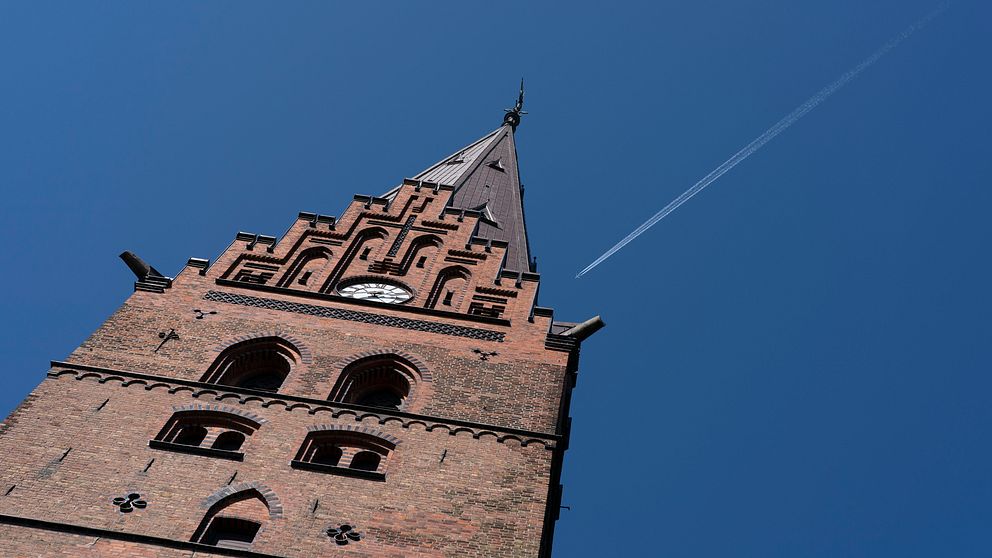 Tornet på en kyrka reser sig mot en klar blå himmel, med en flygplansådra som skär genom luften. Klocktornet av tegel har detaljerade fönster och en spira på toppen.