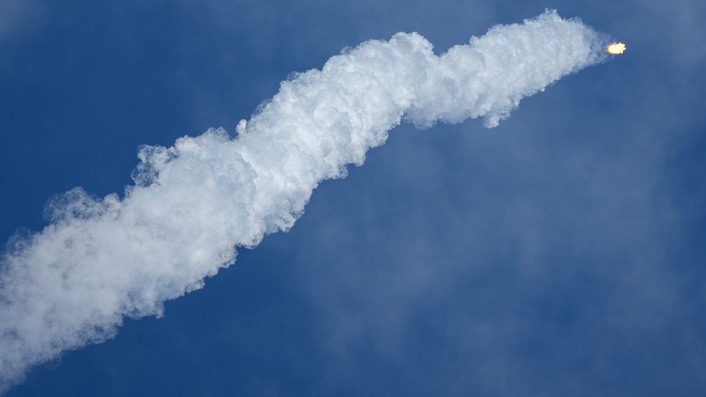 Rök och eldstråle från en raket som skjuts upp mot en klarblå himmel.